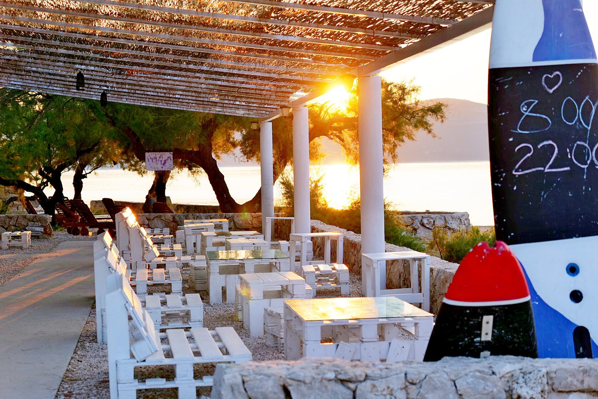 Sunset view of the café at the sustainable Hotel Korinjak with white pallet furniture and a pergola. Sunlight filters through the trees at the water's edge, creating a serene atmosphere.