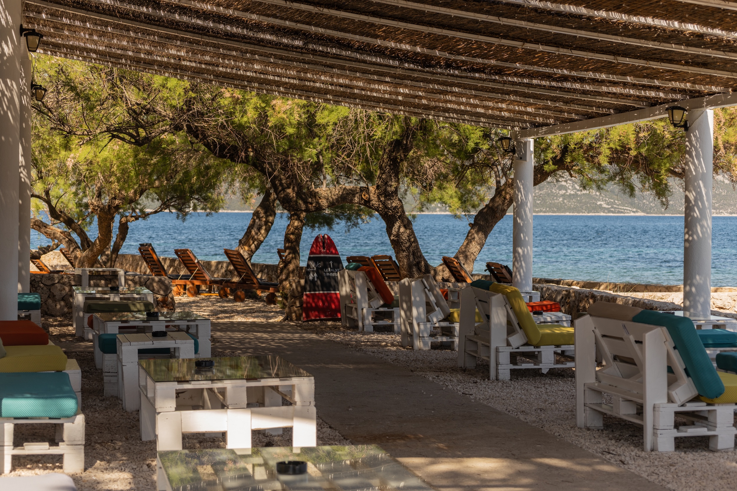 Überdachte Strandbar mit weißen Holzmöbeln und bunten Polstern vor Bäumen und Meer im Hotel Korinjak in Kroatien