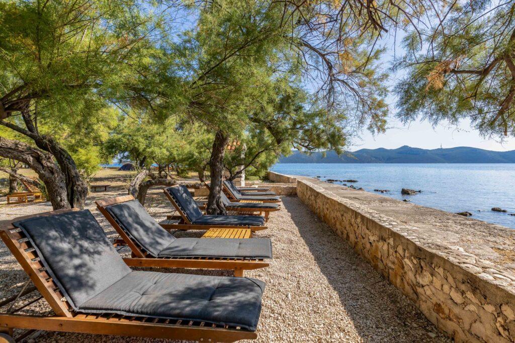 Mehrere Liegestühle mit dunkelgrauen Polstern stehen unter Bäumen auf einem Kiesstrand neben einer niedrigen Steinmauer am Meer im Hotel Korinjak in Kroatien