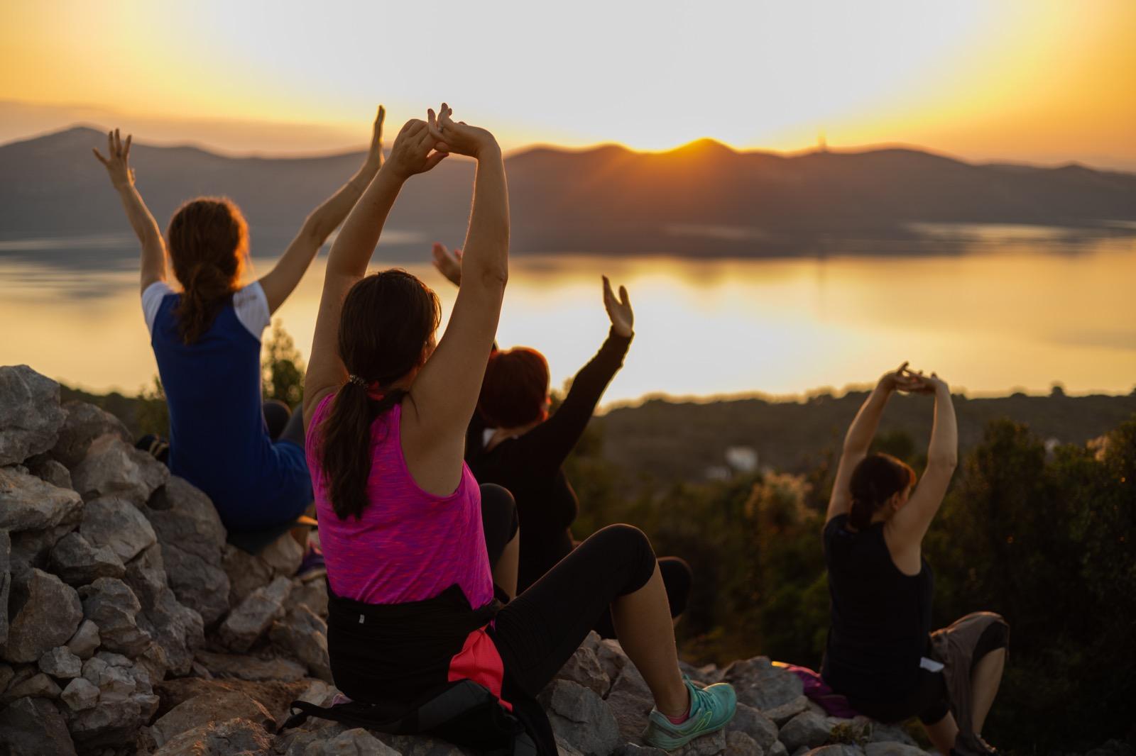 Four people are sitting on a rock at sunset, their arms raised in a yoga pose. The lake and mountains in the background create a peaceful atmosphere.