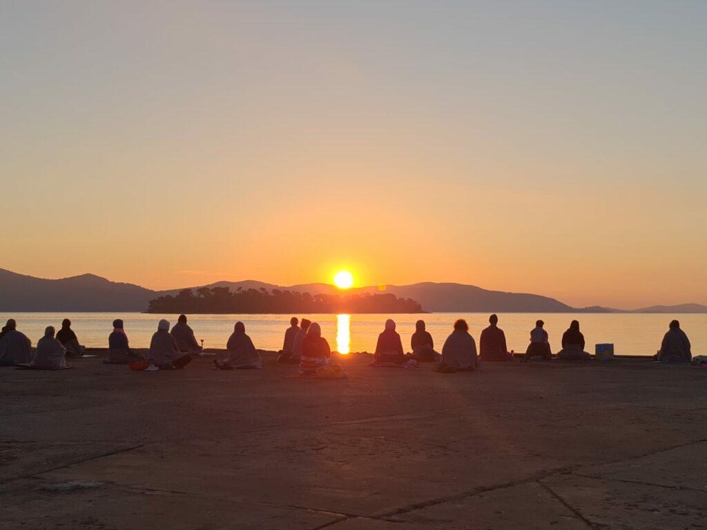 Eine Gruppe von Menschen sitzt im Schneidersitz auf einem Pier nahe des nachhaltigen Hotel Korinjak und blickt bei Sonnenaufgang auf das ruhige Meer. Der Himmel ist in Orange- und Rosatöne getaucht, was eine friedliche Atmosphäre schafft.