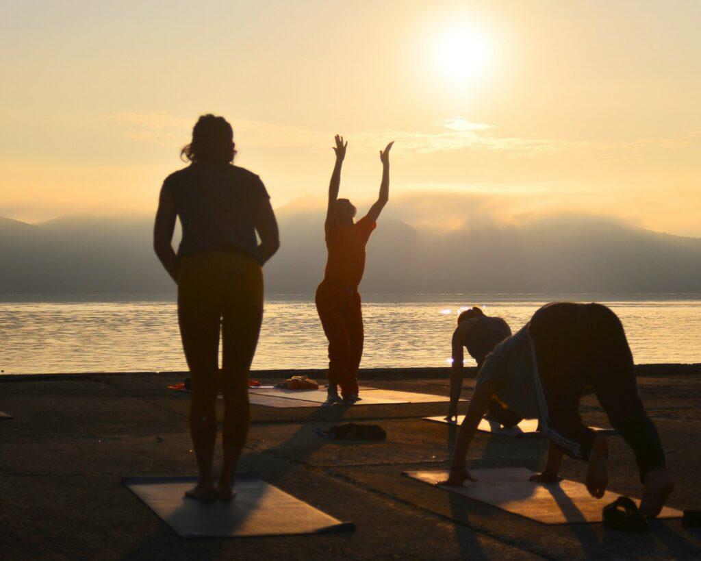 Menschen in Silhouetten praktizieren bei Sonnenaufgang Yoga an einer ruhigen Küste vor dem nachhaltigen, vegetarischen Hotel Korinjak in Kroatien. Eine Person steht mit erhobenen Armen da und strahlt Ruhe und Konzentration aus.
