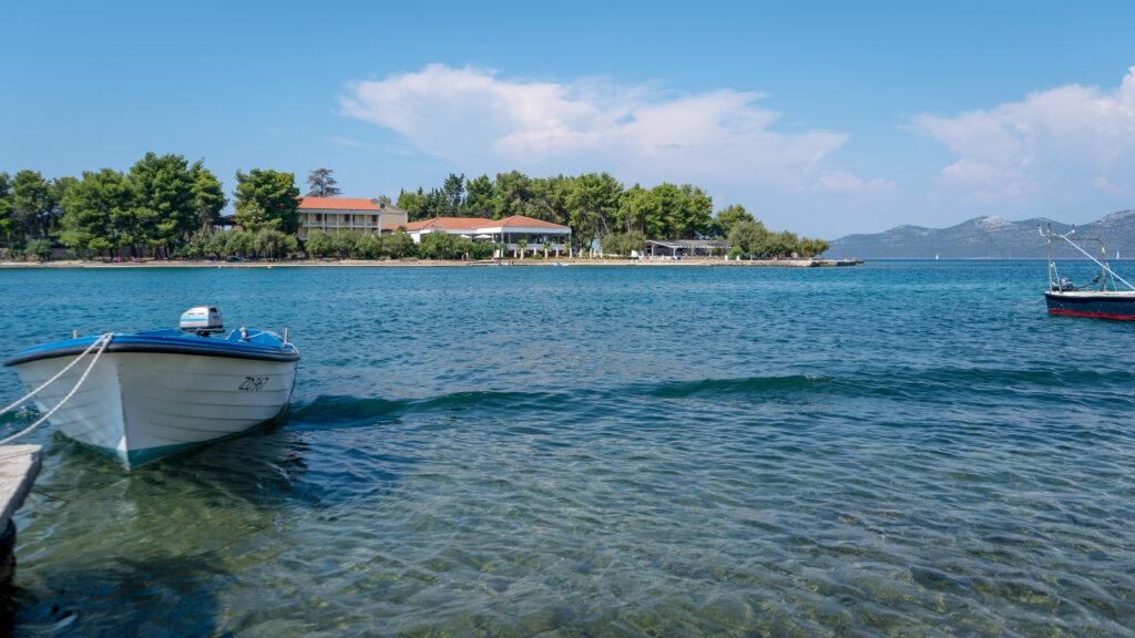 Ruhiges türkisfarbenes Meer mit kleinen Booten, die in der Nähe der üppigen, baumbestandenen Insel Iz in Kroatien anlegen. Ein klarer blauer Himmel, die rustikalen Gebäude des nachhaltigen Hotel Korinjak und ferne Hügel schaffen eine ruhige, malerische Szene.