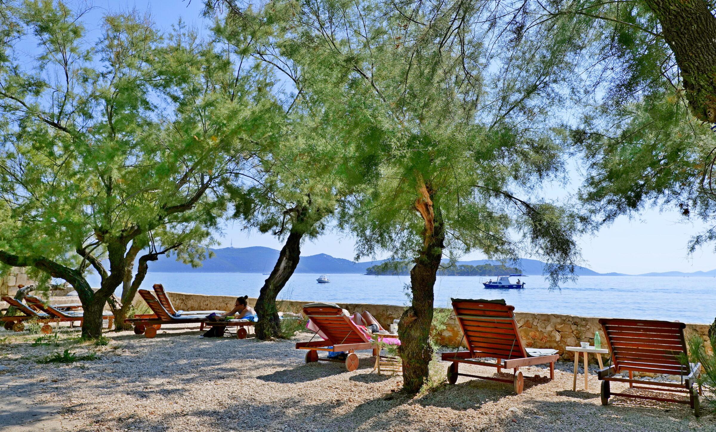 Sun-drenched beach scene at the Korinjak Hotel in Croatia, with wooden deckchairs under green trees, looking out to the calm sea. In the distance, boats bob up and down on the waves, creating a relaxed, tranquil atmosphere.