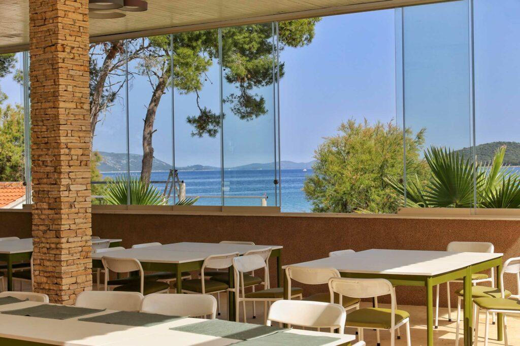 Essbereich des nachhaltigen Hotel Korinjak auf der Insel Iz, Kroatien mit weißen Tischen und Stühlen unter einer Holzdecke. Eine Steinsäule und eine Glaswand geben einen ruhigen Blick auf das Meer und die Bäume frei.