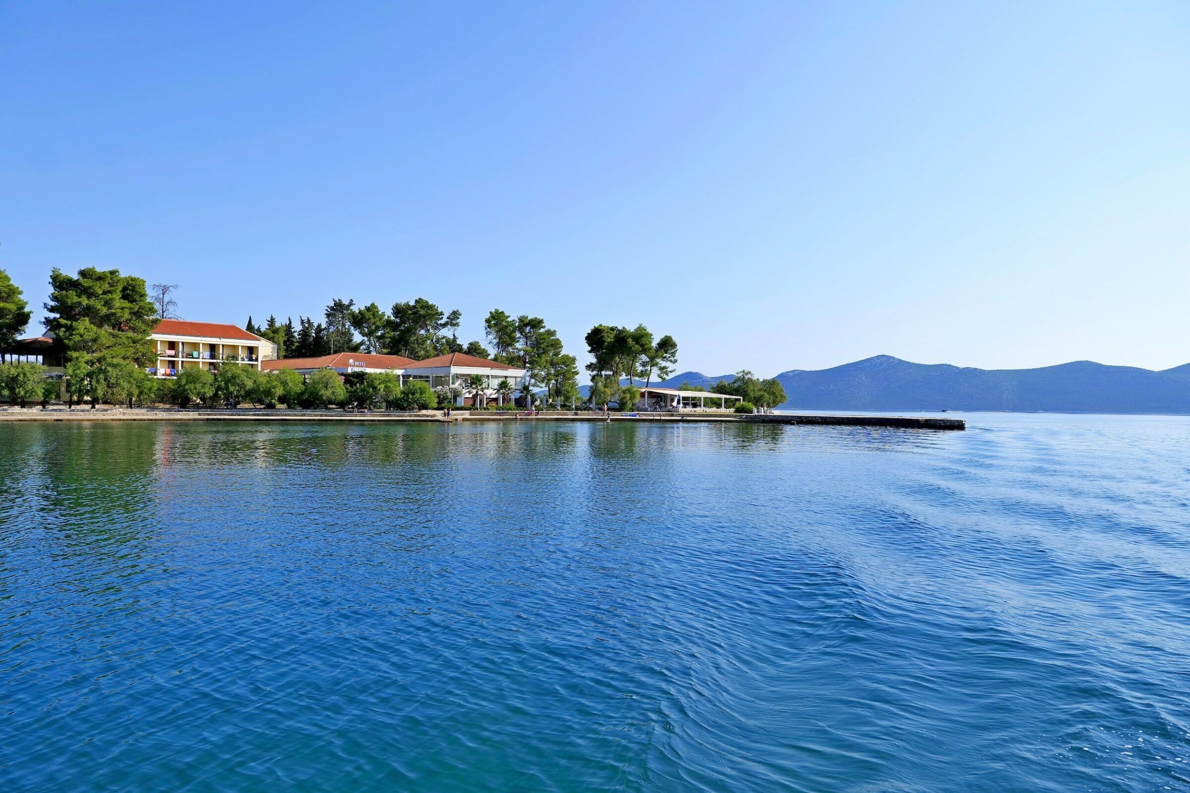 Blick auf das kroatische Hotel Korinjak und auf das ruhige blaue Meer mit sanften Wellen, einer Küstenlinie mit Gebäuden und Bäumen unter einem klaren Himmel und fernen Bergen am Horizont.