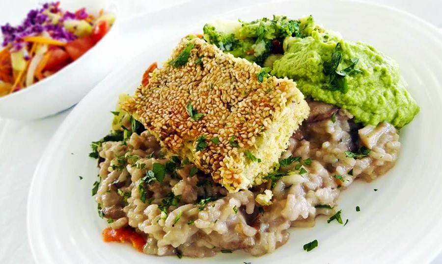 Ein farbenfrohes vegetarisches Gericht mit Tofu in Sesamkruste, cremigem Risotto und grünem Gemüsepüree, garniert mit Kräutern. Im Hintergrund ist eine bunte Salatschüssel zu sehen. Im nachhaltigen Hotel Korinjak