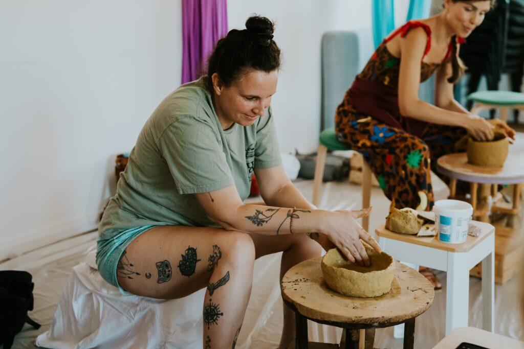 Zwei Frauen sitzen an Töpferscheiben und formen Ton, die vordere trägt ein grünes T-Shirt und Shorts mit sichtbaren Tattoos an Armen und Beinen