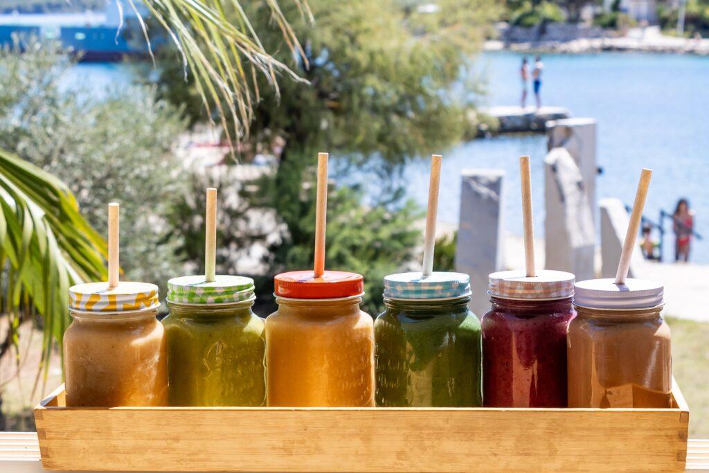 Sieben bunte Smoothies in Einmachgläsern mit Strohhalmen stehen auf einem Holztablett vor einer Wasserlandschaft mit Palmen und Menschen im Hintergrund.