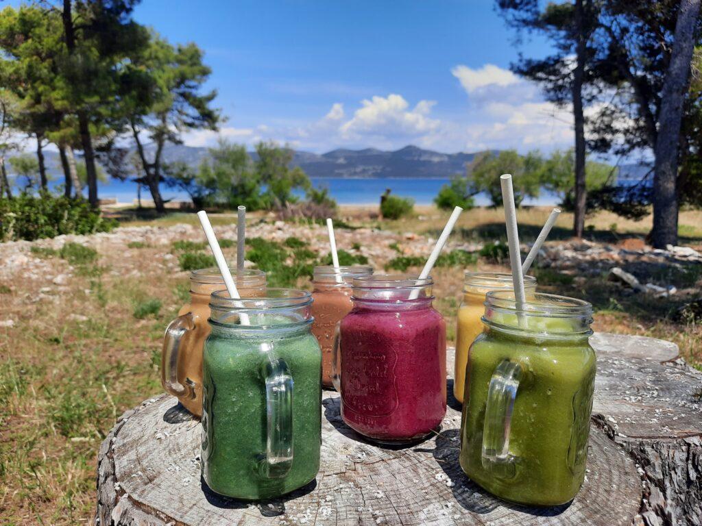 Eine Vielzahl bunter Smoothies in Einmachgläsern mit Strohhalmen auf einem Baumstumpf, vor der malerischen Kulisse der kroatischen Insel Iz aus Bäumen, einem ruhigen Meer und Bergen. Vor dem nachhaltigen Hotel Korinjak