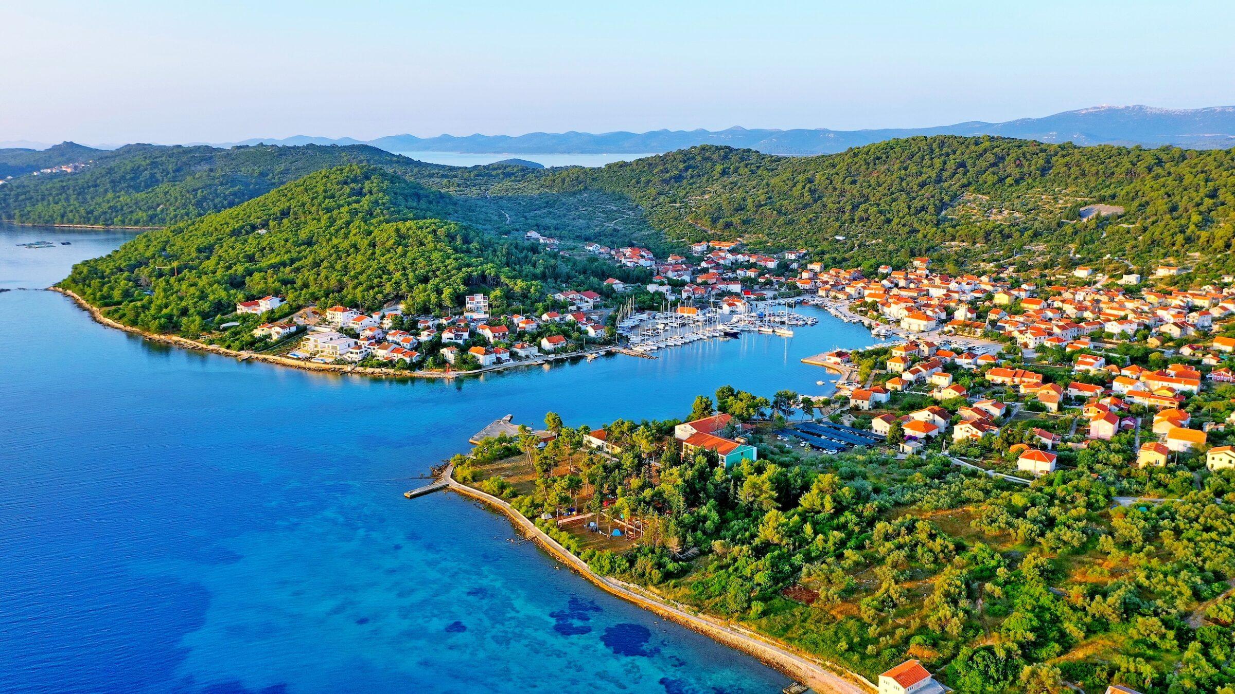 Luftaufnahme eines beschaulichen Küstendorfes auf der kroatischen Insel Iz mit leuchtend roten Dächern, üppig grünen Hügeln und einem Jachthafen voller Segelboote unter einem klaren blauen Himmel.