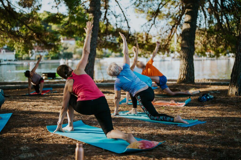 Mehrere Personen praktizieren Yoga auf blauen Matten im Freien unter Bäumen am Ufer eines Gewässers im Hotel Korinjak in Kroatien.