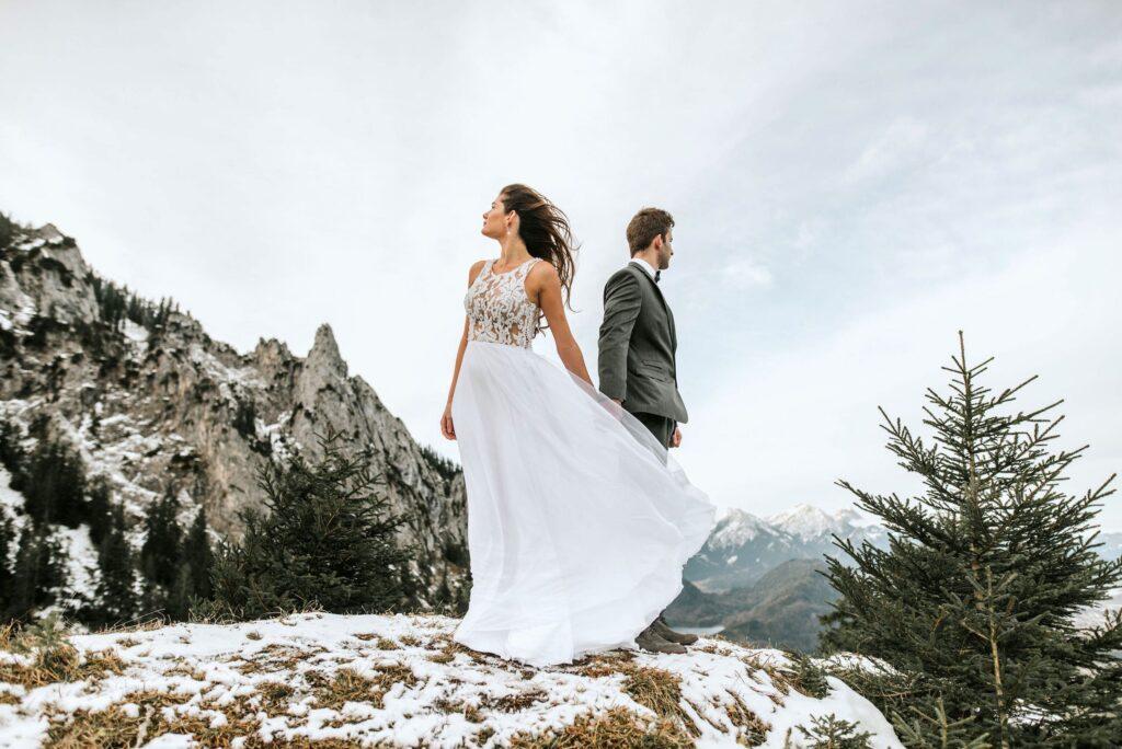 Eine Braut in einem wallenden weißen Kleid und ein Bräutigam im Anzug stehen Rücken an Rücken auf einem verschneiten Berggipfel in der Nähe des nachhaltigen Hotels Das Rübezahl in Bayern und vermitteln ein Gefühl von Abenteuer und Romantik.