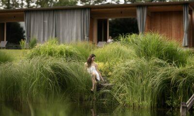 Eine Frau in einem weißen Bademantel sitzt friedlich zwischen hohen grünen Gräsern an einem ruhigen Teich. Im Hintergrund ist das Holzgebäude der Gartenblickzimmer des STURM mit offenen Vorhängen zu sehen.