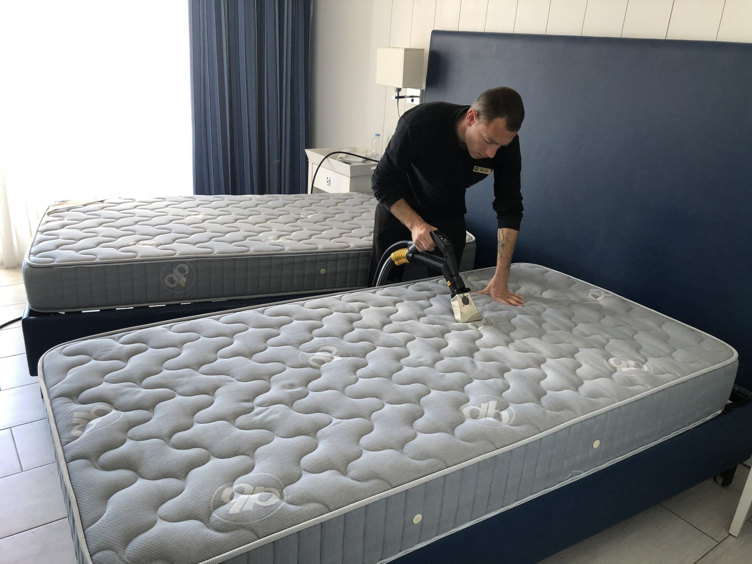 Person, die eine Matratze mit einem Microtrockendampfreiniger von medeco Cleantec in einem hellen Hotelzimmer reinigt. Zwei Einzelbetten mit grau gemusterten Bezügen. Die Atmosphäre ist professionell und ordentlich.