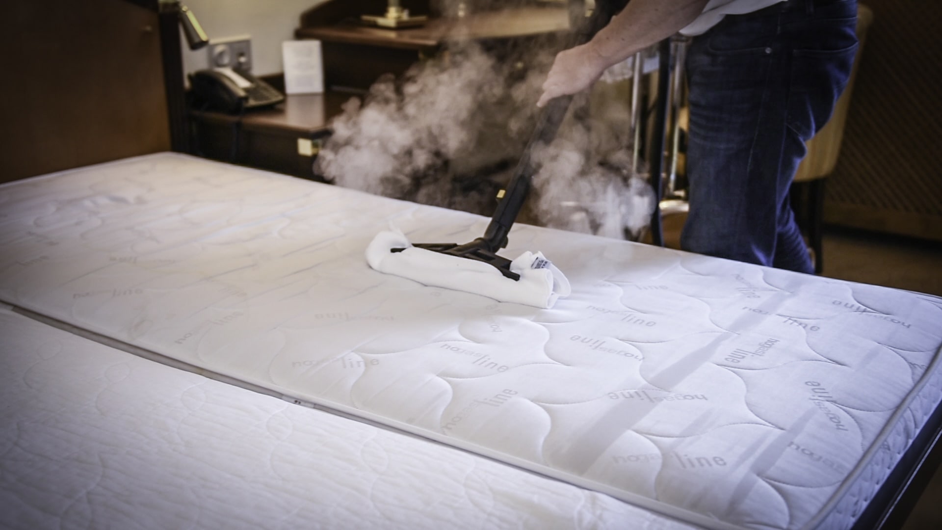 Eine Person reinigt eine Matratze in einem Zimmer mit Microtrockendampf, wobei sichtbarer Dampf aufsteigt. Das Bett ist ordentlich gemacht, wodurch eine saubere und hygienische Atmosphäre entsteht.