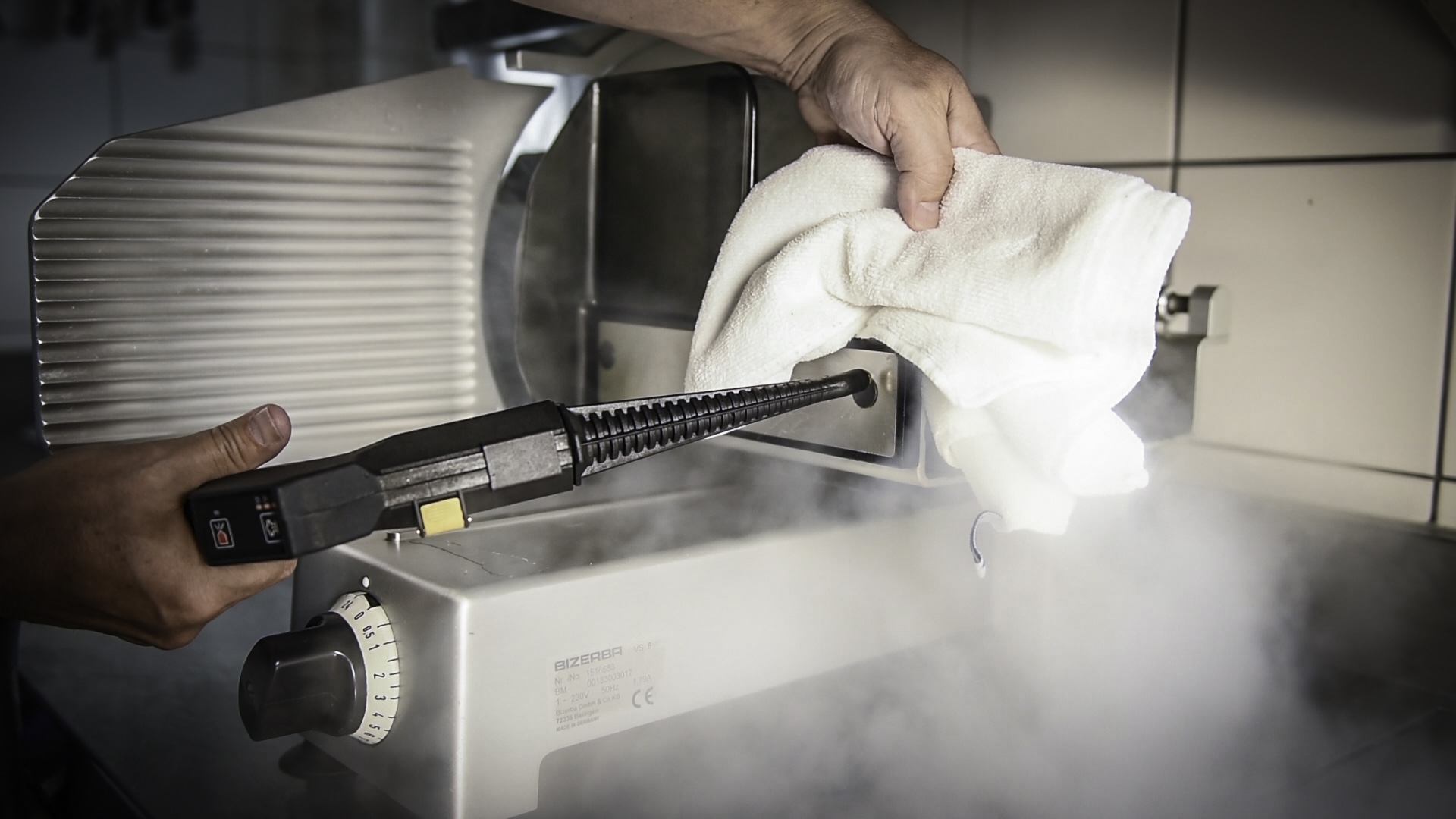 Eine Person benutzt einen Microtrockendampfreiniger von Medeco Cleantec für ein Küchengerät und hält dabei ein Tuch zum Abwischen bereit. Der Dampf vernebelt die Szene und suggeriert Sauberkeit und Hygiene.