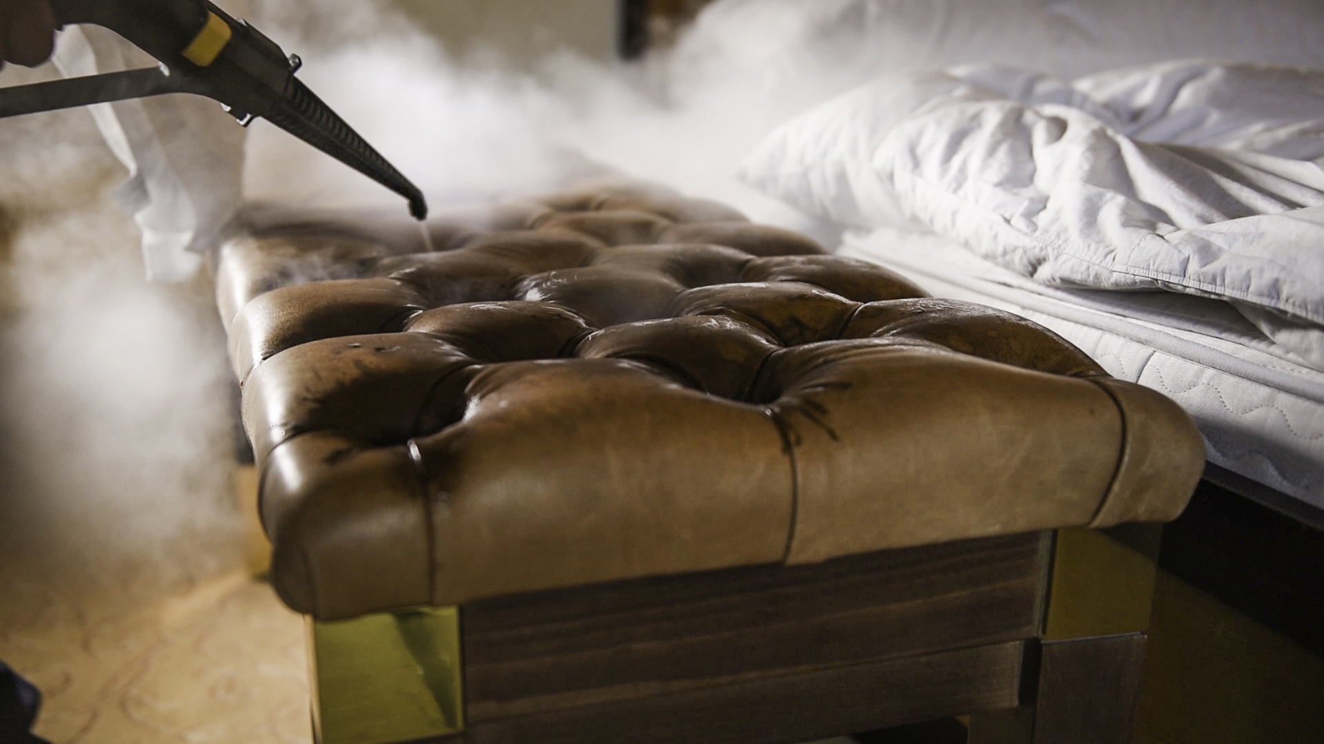 Ein Microtrockendampfreiniger wird auf einer braunen Lederbank neben einem Bett mit weißen Laken verwendet. Der Dampf erzeugt einen nebligen Effekt, der Sauberkeit und Sorgfalt unterstreicht.
