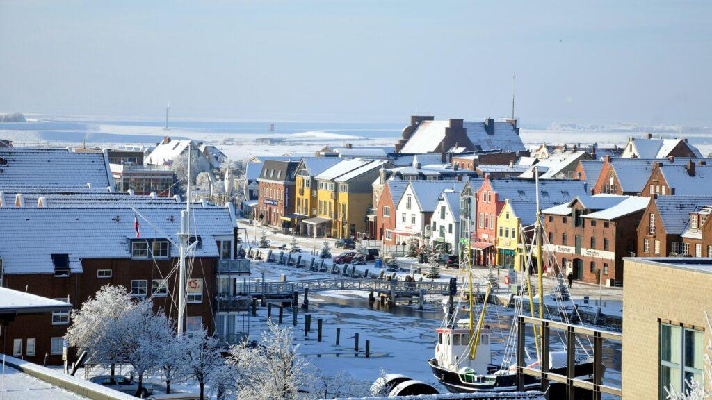 The snow-covered port town of Husum with its colorful buildings lines a quiet waterfront promenade. Snow-covered roofs and boats create a peaceful winter landscape under clear skies.