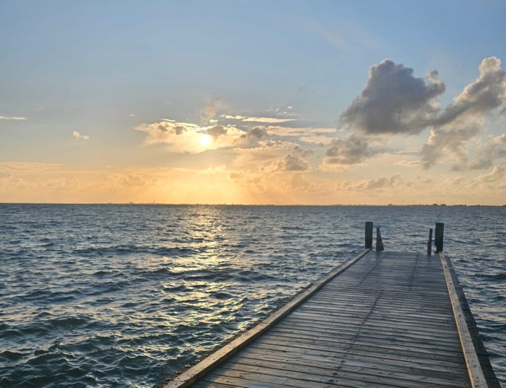A wooden walkway juts out into the calm North Sea near Husum under a glowing sunset sky. Fluffy clouds and golden sunlight reflect on the water, creating a peaceful, tranquil atmosphere.
