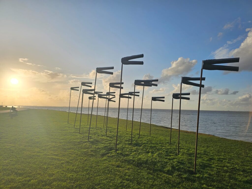 Sunset over a tranquil coastline with modern wind sculptures on a grassy area. The sky is partly cloudy, creating a peaceful and picturesque scene.