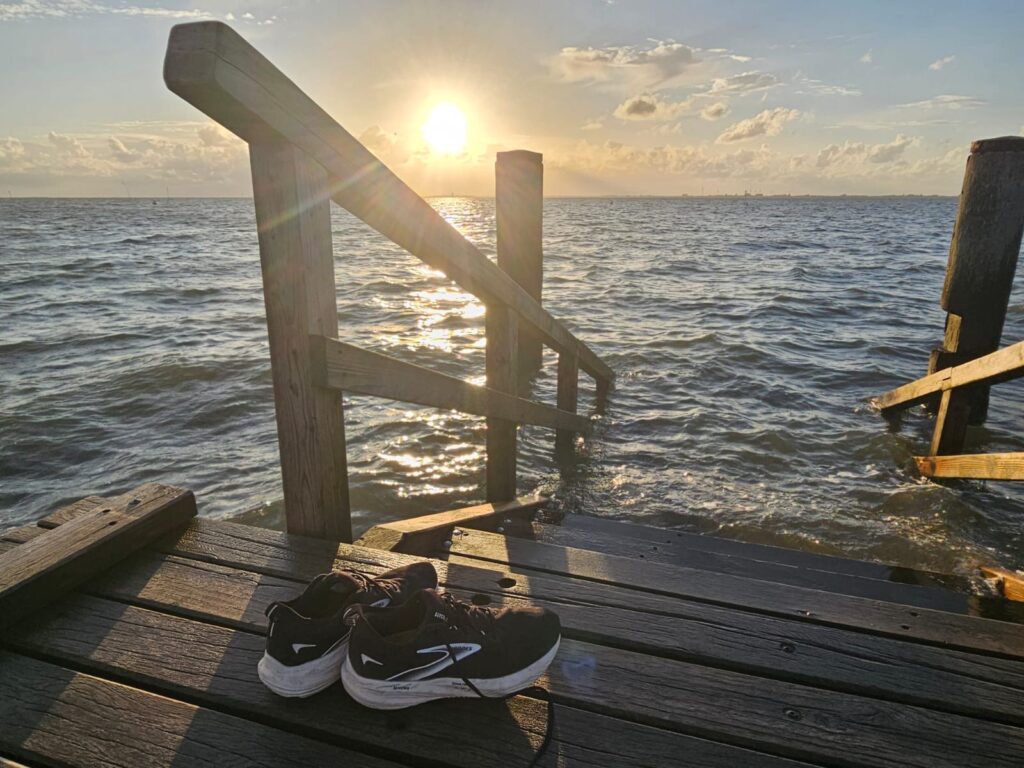 A pair of black sneakers stands on a wooden deck overlooking the North Sea near Husum. A sunlit staircase leads down into the water as the sun sets on the horizon.