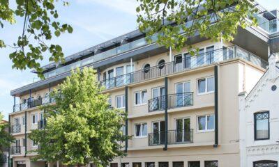 Modern four-story apartment building of the sustainable boutique hotel SANDglow in Husum with balconies, surrounded by green trees under a bright blue sky, conveying an urban and peaceful atmosphere.