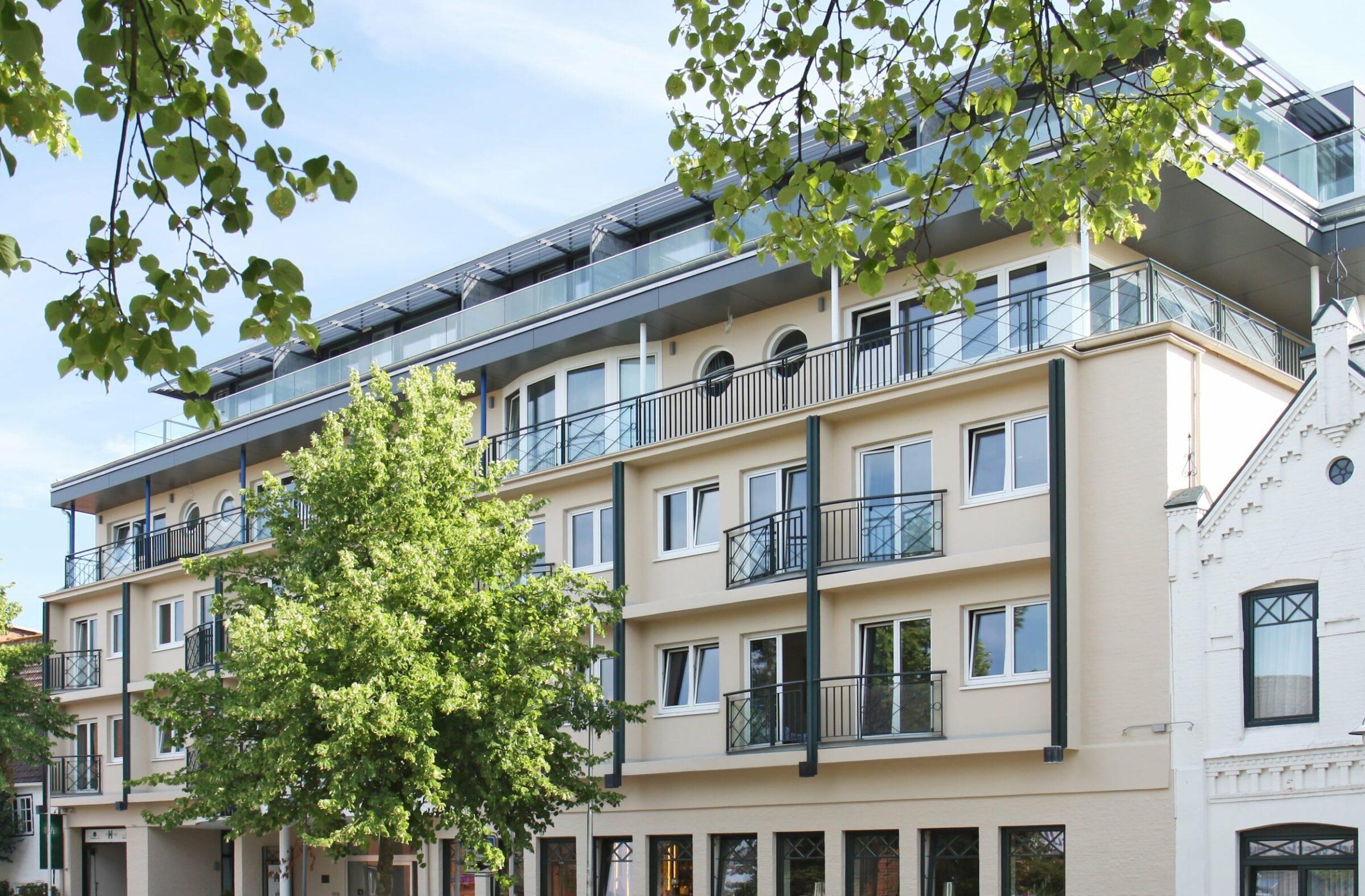 Modern four-story apartment building of the sustainable boutique hotel SANDglow in Husum with balconies, surrounded by green trees under a bright blue sky, conveying an urban and peaceful atmosphere.