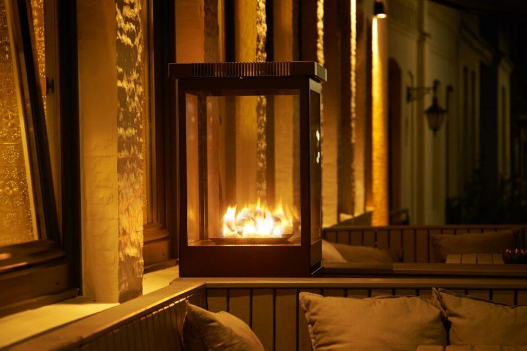Cozy outdoor terrace at the green boutique hotel SANDglow in Husum with a modern glass fireplace that gives off a warm glow. Soft cushions line the wooden seats and create a welcoming, peaceful evening atmosphere.