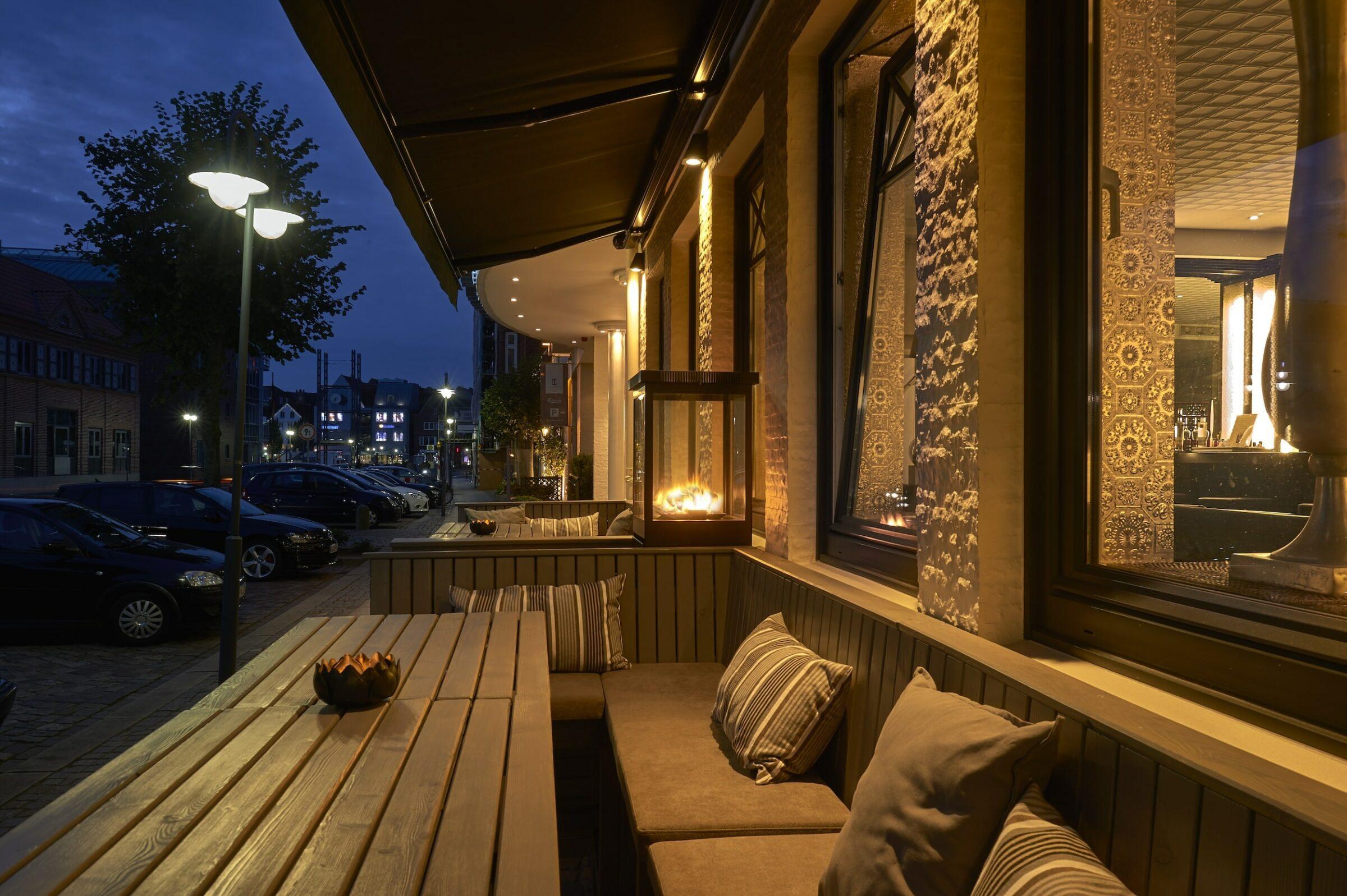 Comfortable outdoor seating in front of SANDglow, a sustainable adults-only hotel in Husum, at night with a long wooden table, upholstered benches, and warm lighting. A small fire illuminates the atmospheric space.