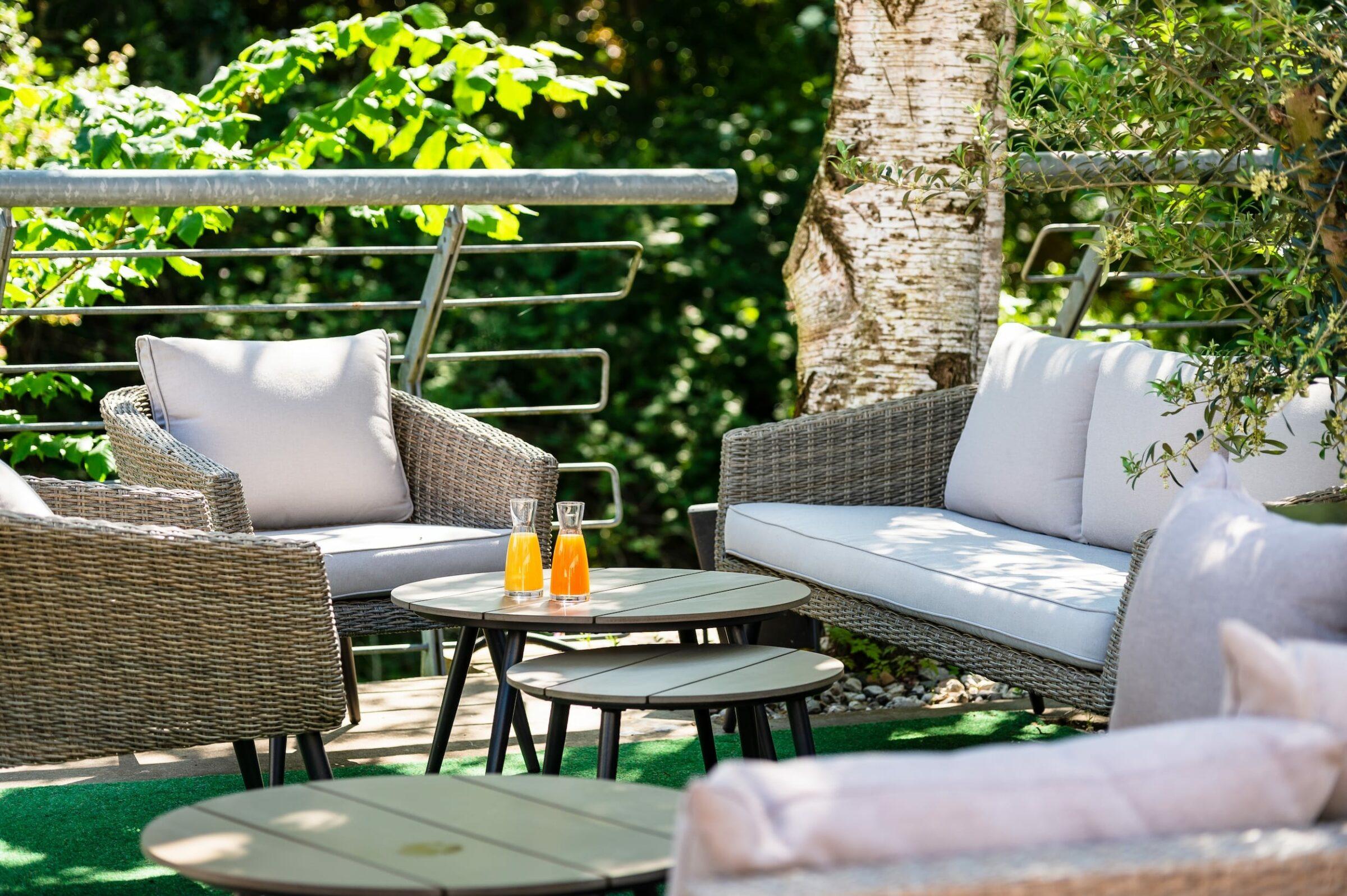 Outdoor area of the sustainable boutique hotel SANDglow in Husum, northern Germany, with wicker chairs and a sofa with cushions, surrounded by lush greenery. Two glasses of orange juice on a table invite you to relax.