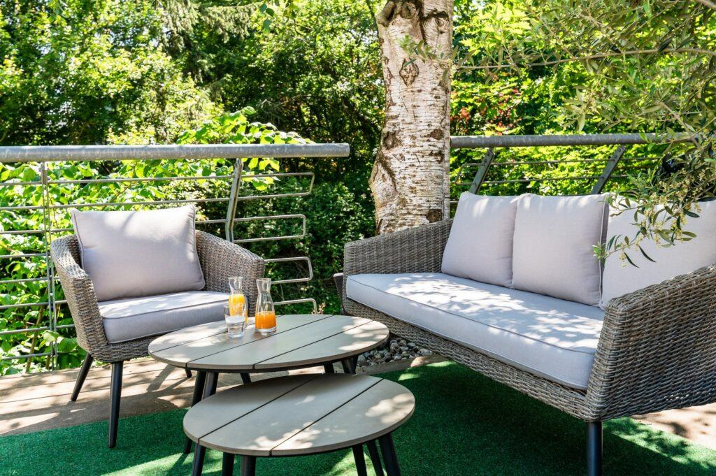 Outdoor patio with rattan furniture, including an upholstered two-seater sofa and an armchair, at the sustainable SANDglow business hotel in Husum, which is exclusively for adults. Glasses of orange juice stand on two round tables. Lush greenery and trees create a peaceful atmosphere.