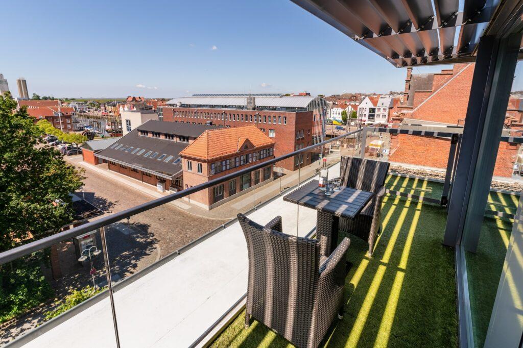 Roof terrace of the modern and sustainable SANDglow hotel with wicker chairs and a table, overlooking the picturesque harbor town of Husum. Warm sunlight casts shadows on the grass floor and creates a peaceful, relaxed atmosphere.
