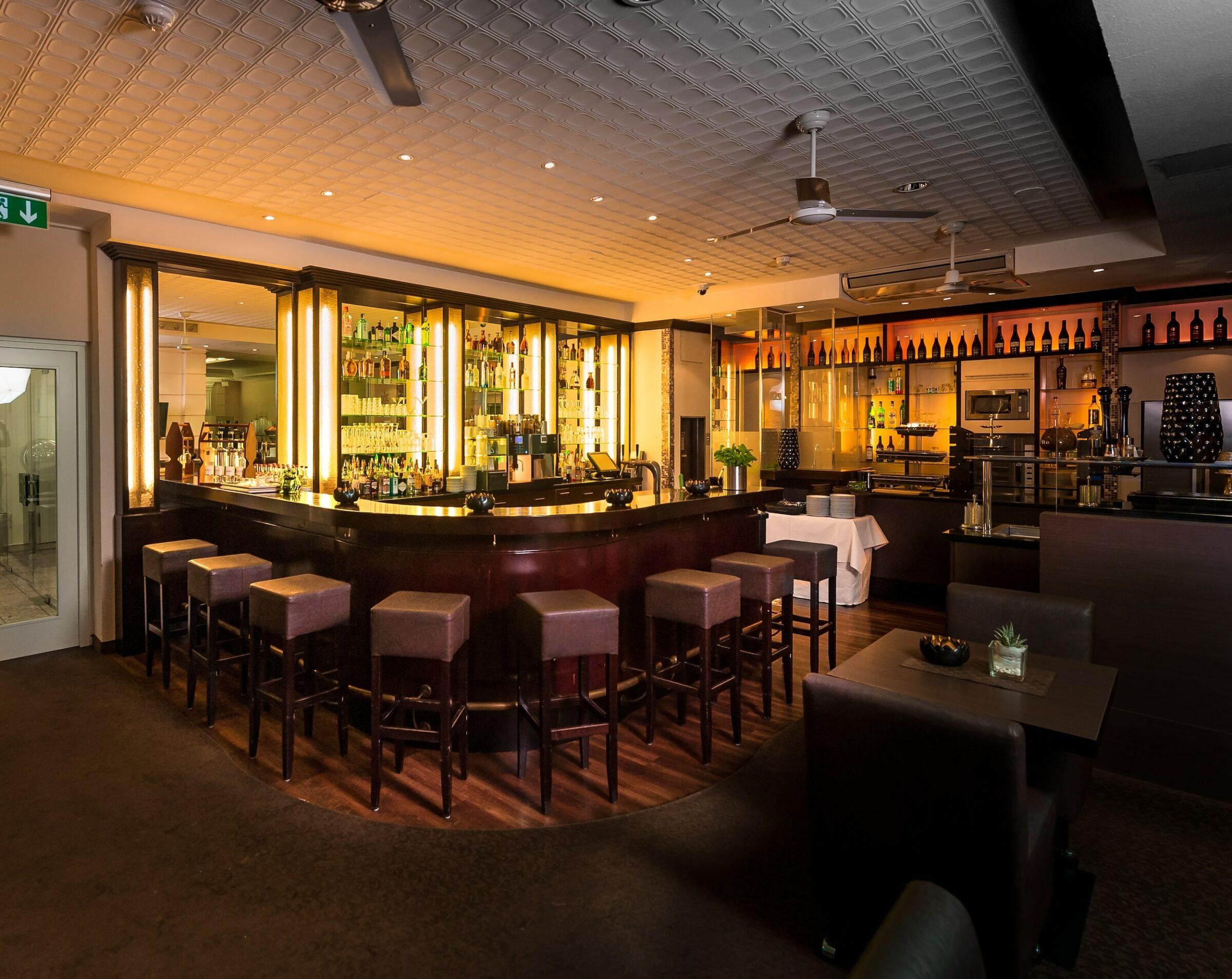 Elegant bar interior with a warm, cozy atmosphere in the modern and cosmopolitan eco-boutique hotel SANDglow. Featuring an elegant, curved bar with high stools, shelves lined with bottles, and soft lighting.