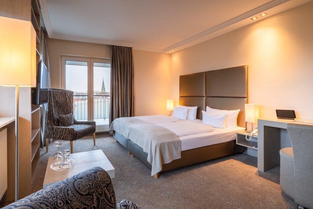 Hotel room at the eco-friendly and chic SANDglow Hotel with modern, cozy design, double bed, elegant armchair, soft lighting, and a window overlooking the city and a distant tower.
