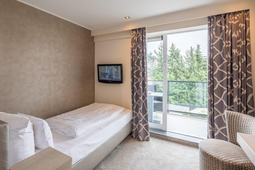 Cozy hotel room with a single bed, wall-mounted TV, patterned curtains, and a chair at the sustainable SANDglow business hotel in Husum on the North Sea coast. Glass doors lead to a balcony with a view of lush greenery.