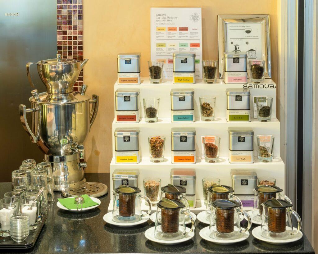 A tea display with various tea caddies on white shelves, each paired with transparent cups containing loose tea leaves, can be found at the fair breakfast buffet of the eco-hotel SANDglow. A large metal samovar and several glass cups stand on the counter, creating a warm, inviting atmosphere.