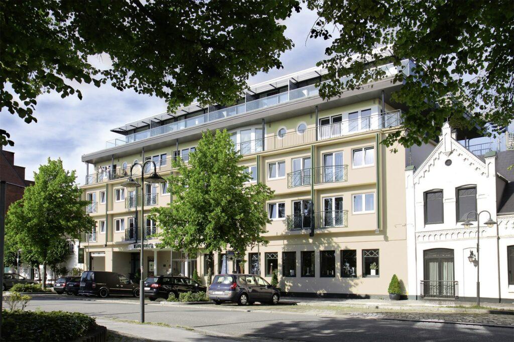 Street view of the modern, beige, four-story boutique hotel SANDglow with balconies, flanked by trees and parked cars. Adjacent is a white building in historic style.