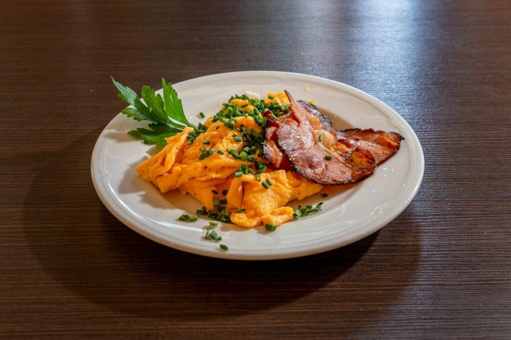 A plate of freshly prepared scrambled eggs, garnished with chopped chives and crispy bacon slices, decorated with parsley on a wooden table—a fresh, inviting breakfast scene from the eco-boutique hotel SANDglow.