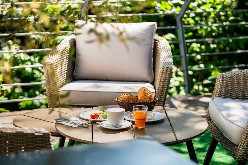 Outdoor patio at the adults-only SANDglow hotel with a comfortable wicker chair, a table with breakfast consisting of coffee, orange juice, salad, and bread, surrounded by lush greenery.