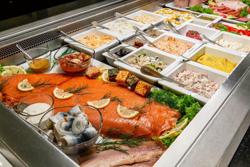 Regional and fair trade breakfast buffet at the eco-friendly Hotel SANDglow, featuring smoked salmon slices garnished with lemon and dill, accompanied by a variety of salads, sauces, and marinated fish in a refrigerated display case.