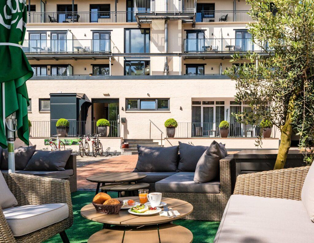 Outdoor hotel terrace with wicker furniture and tables for breakfast, including croissants and juice. Modern building facade of the sustainable SANDglow Hotel with balconies in the background.