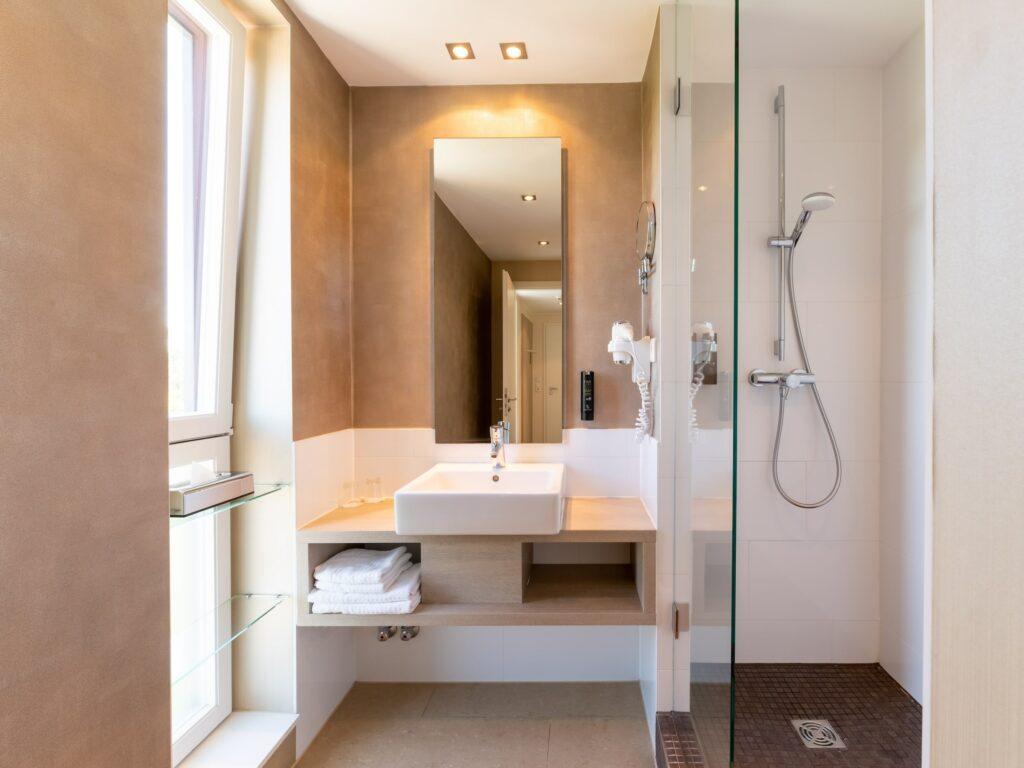Modern bathroom interior with an elegant white sink, a large mirror, and a glass shower in the chic and sustainable boutique hotel SANDglow in Husum. Natural light streams in through a window, emphasizing the clean, minimalist design.