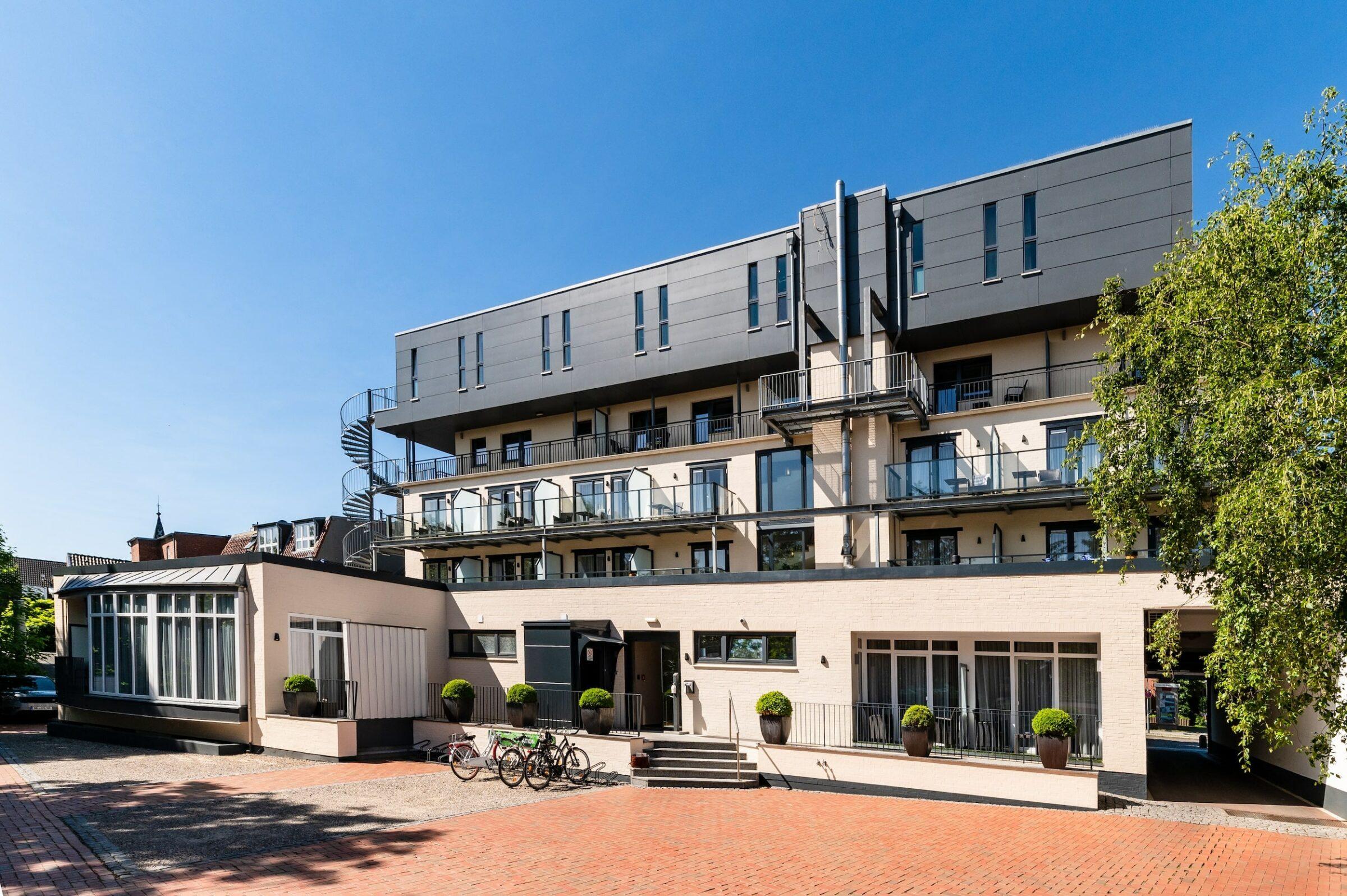 A modern four-story building with balconies and large windows: the sustainable SANDglow hotel for adults only. A spiral staircase connects the upper floors. Outside, there are myBoo bicycles. Lush trees frame the bright scenery under a bright blue sky.