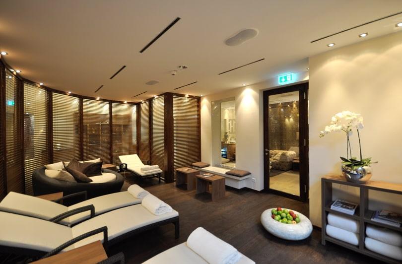 Luxurious spa lounge in the sustainable boutique hotel SANDglow on the North Sea coast—with deck chairs, soft lighting, and wooden décor. Rolled towels, a bowl of apples, and a white orchid are arranged on a table, creating a tranquil atmosphere.
