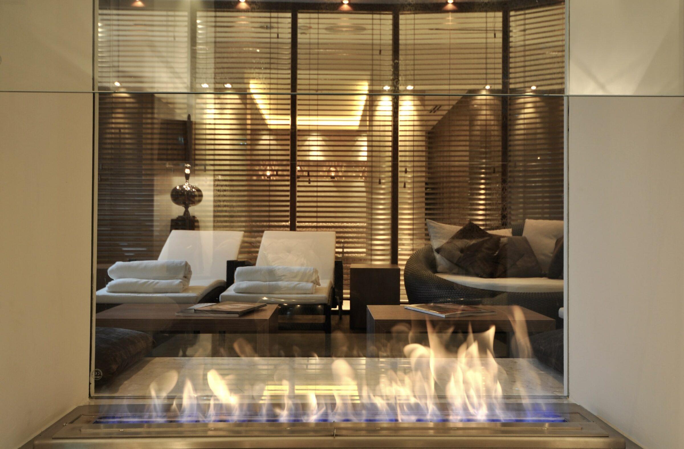 Through a glass wall, you can see a cozy, modern lounge with soft lighting and elegant furniture, with a warm, flickering fire in the foreground. This is the wellness area of the modern boutique hotel SANDglow in the Fairtrade town of Husum.