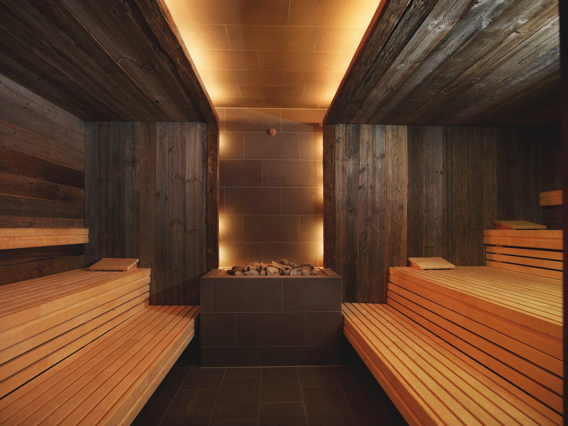 A warm, cozy sauna with rows of wooden benches on both sides, dark wood walls, and a central stove with glowing stones creates a tranquil atmosphere in the wellness area of the sustainable SANDglow hotel in Husum, which is reserved exclusively for adults.