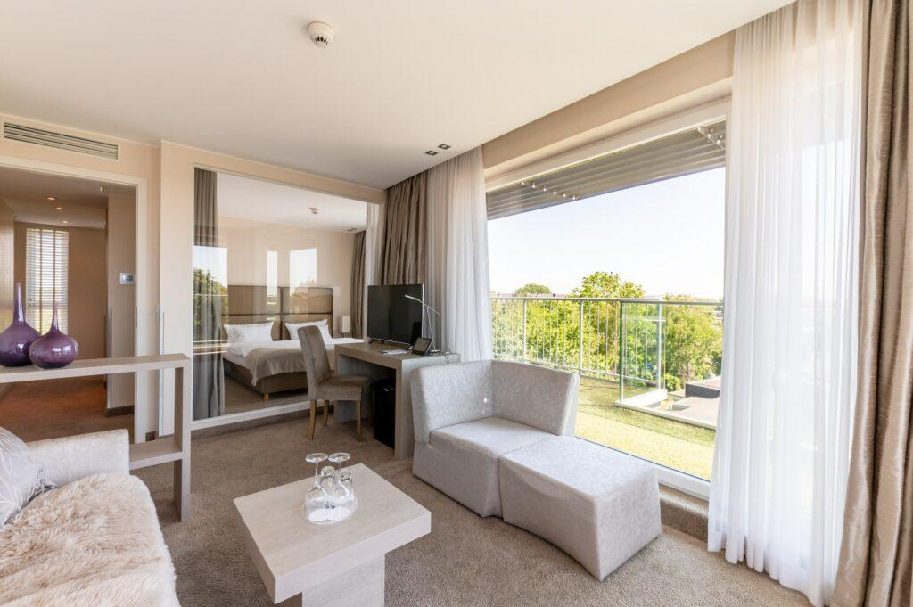 Modern living room in the eco-friendly SANDglow Hotel in Husum, which is reserved for adults only, decorated in beige tones, with a soft sofa, a glass coffee table with two water carafes, and a flat-screen TV. Large windows offer a view of green trees and create a calm, airy atmosphere.