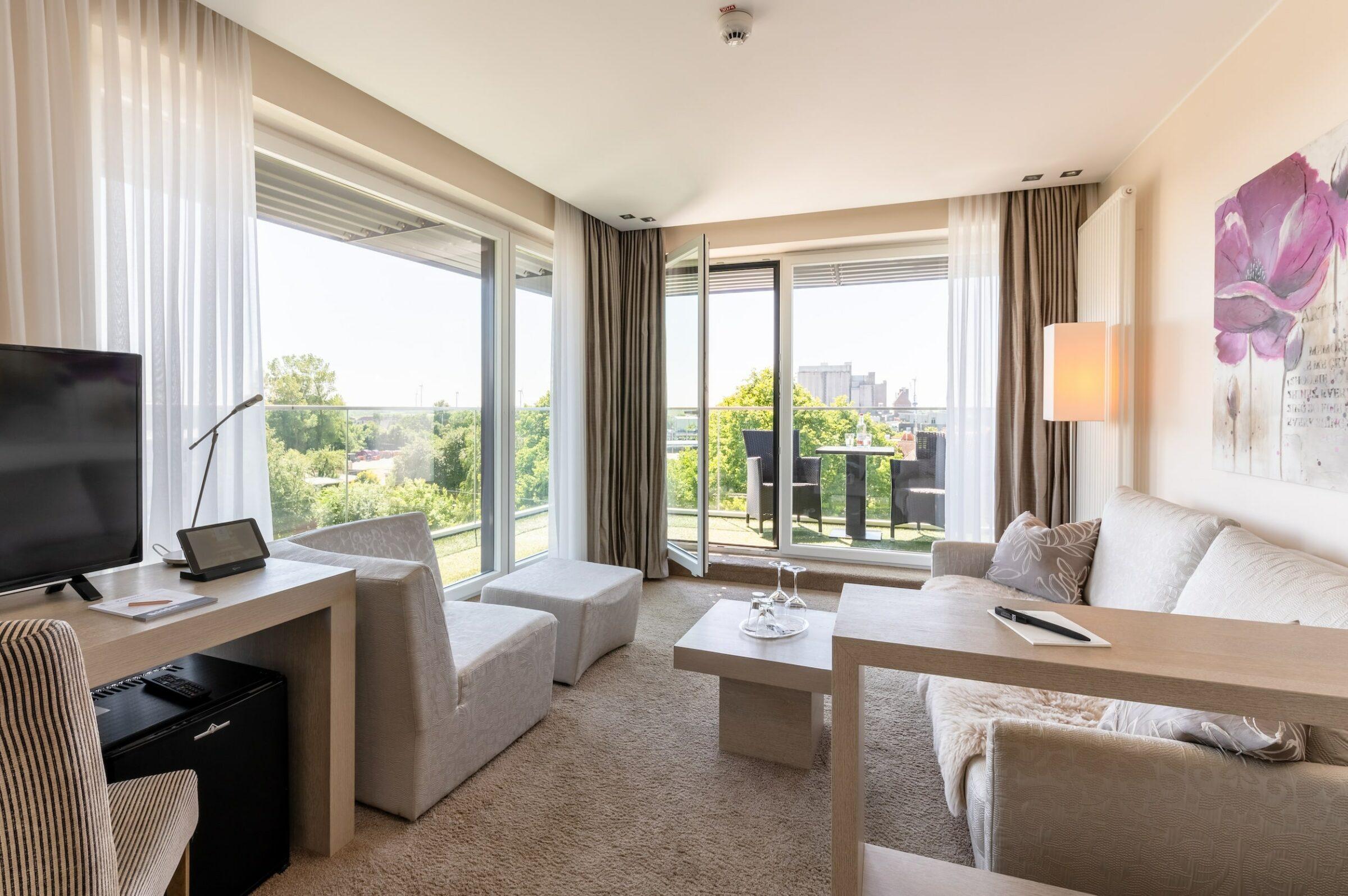 Modern hotel room in the sustainable boutique hotel SANDglow on the North Sea coast—with large windows, light-colored furnishings, and a color palette in shades of beige. The room has a sofa, a desk, a TV, and a balcony with a view of the countryside.