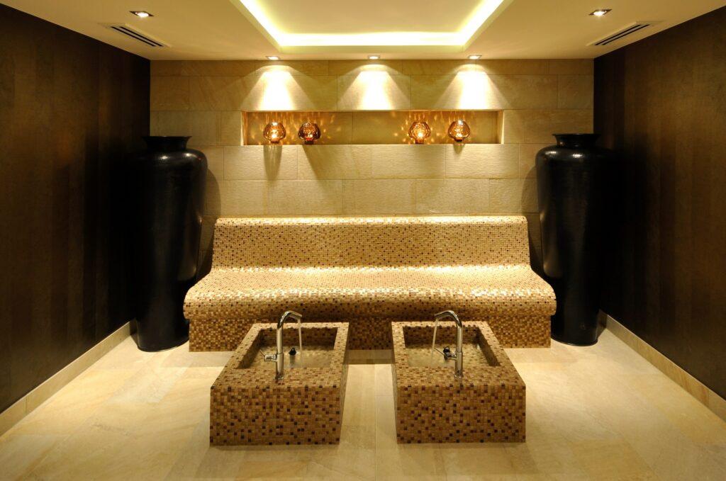 A tranquil wellness room in the sustainable SANDglow hotel on the North Sea coast with soft lighting. A tiled bench stands between large black vases. Two foot basins with taps in front of them create a peaceful atmosphere.